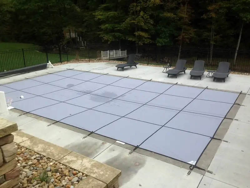 Mesh safety cover stretched tight over an inground pool beside patio loungers