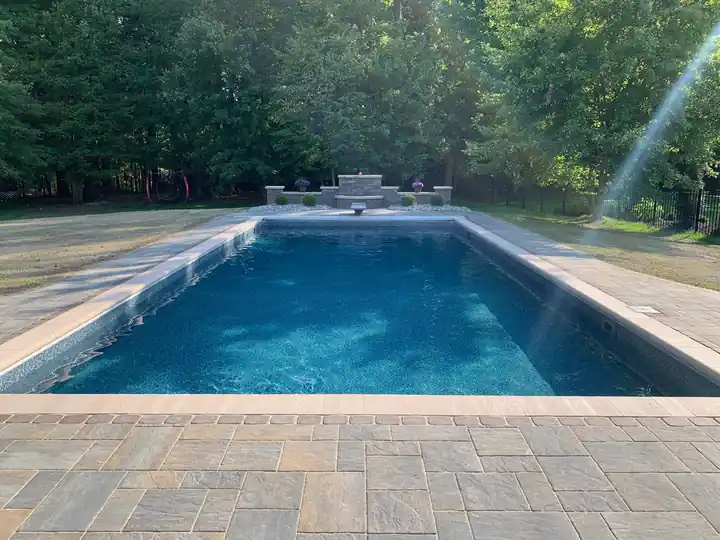 Pool with a paver deck, a stone accent wall and trees behind